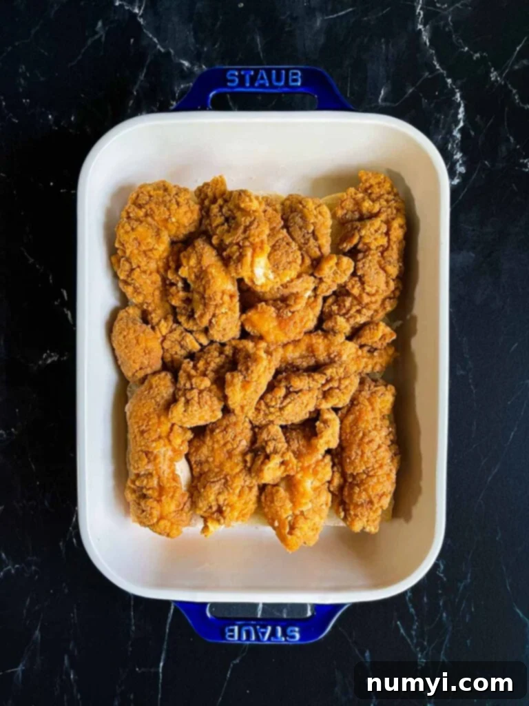 Cooked chicken tenders layered over the bottom halves of slider rolls in a baking dish.