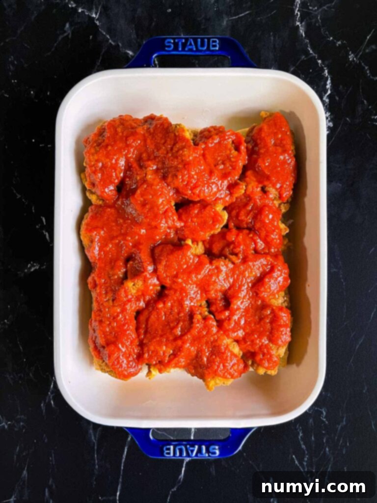 Marinara sauce being spread evenly over the chicken layer on the sliders.
