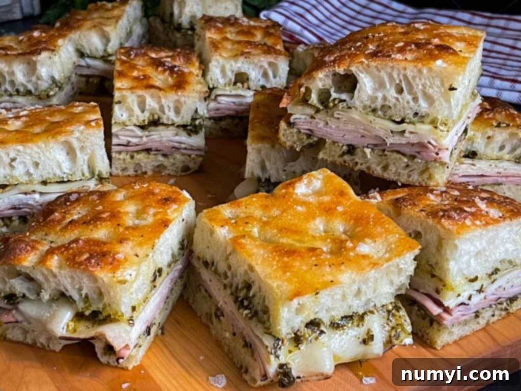 Multiple slices of hot, cheesy pesto focaccia sandwiches with turkey and ham, neatly arranged on a wooden cutting board, ready to be served.