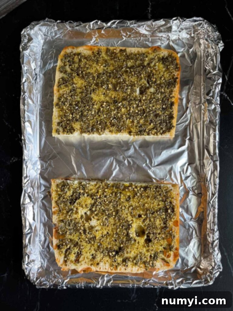 The two halves of focaccia bread, cut side up, with a rich green pesto generously spread over each surface, ready for further layering.