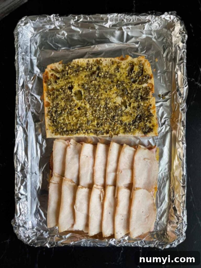Focaccia bread half generously spread with pesto, now topped with layers of thinly sliced turkey, ready for more fillings.