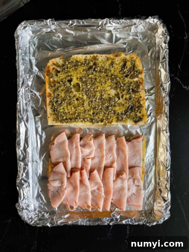 The pesto-spread focaccia, layered with turkey, now receives an even distribution of thinly sliced ham, building a robust sandwich base.