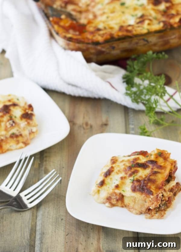 Two plates of freshly cut Five Cheese No-Boil Lasagna, served side by side with forks, and the remaining lasagna in the casserole dish slightly blurred in the background, ready for more servings.
