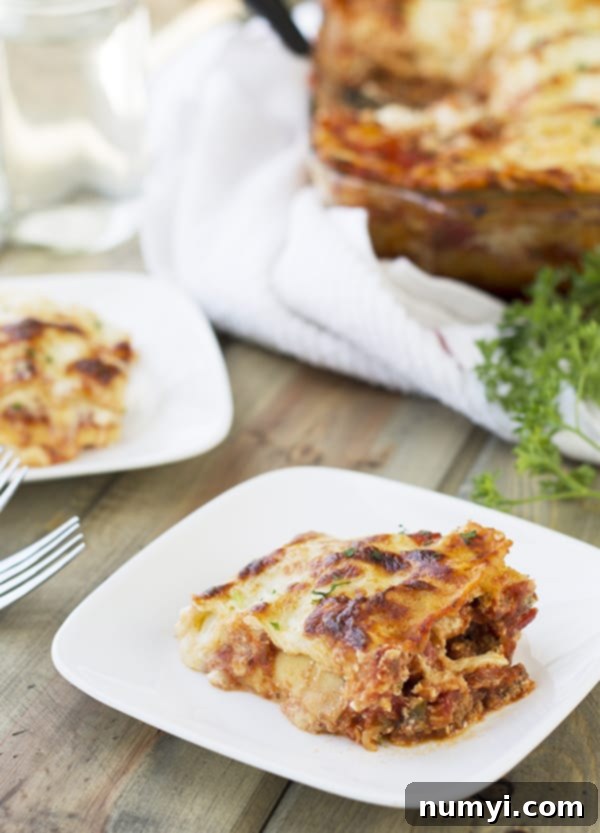 A close-up of a single serving of Five Cheese No-Boil Lasagna on a white plate, with another plate and the full casserole dish blurred in the background, highlighting the golden-brown top and luscious layers.