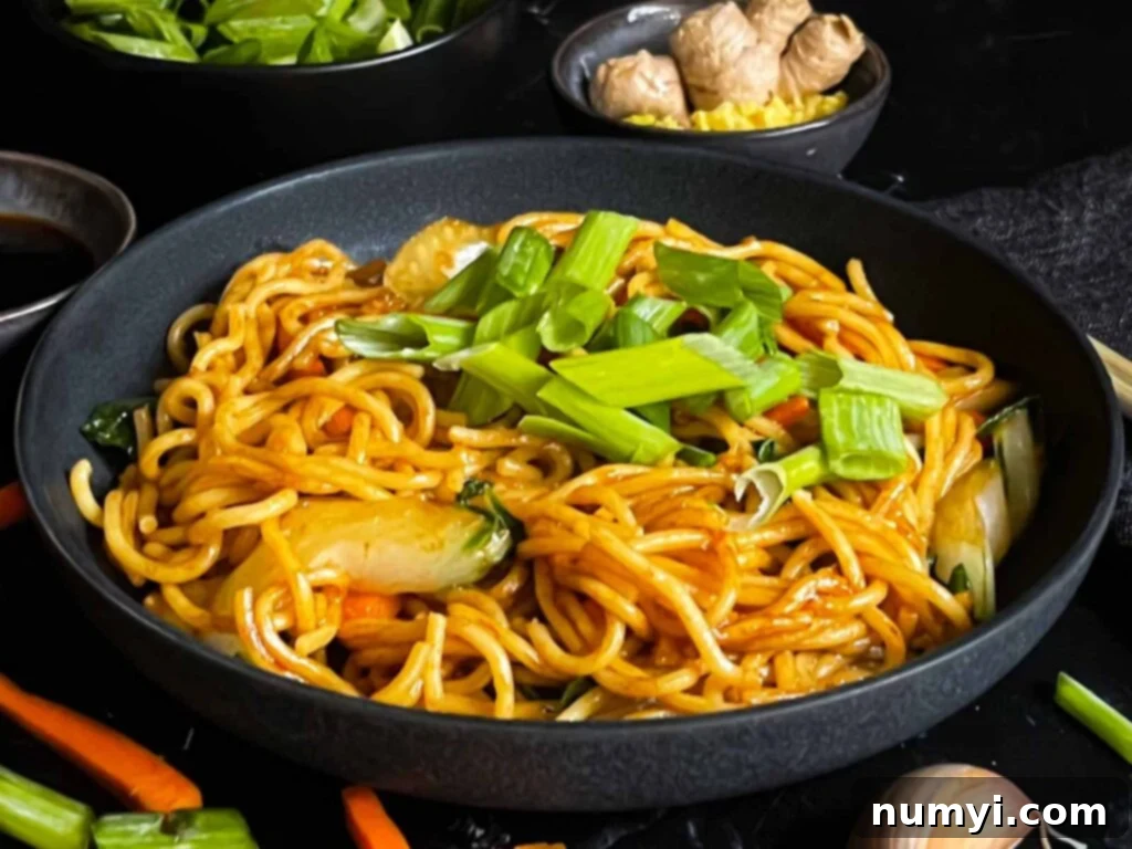 A dark bowl filled with vegetable lo mein, garnished with thinly sliced green onions, with a serving fork ready to highlight the deliciousness of this homemade Chinese noodle dish.