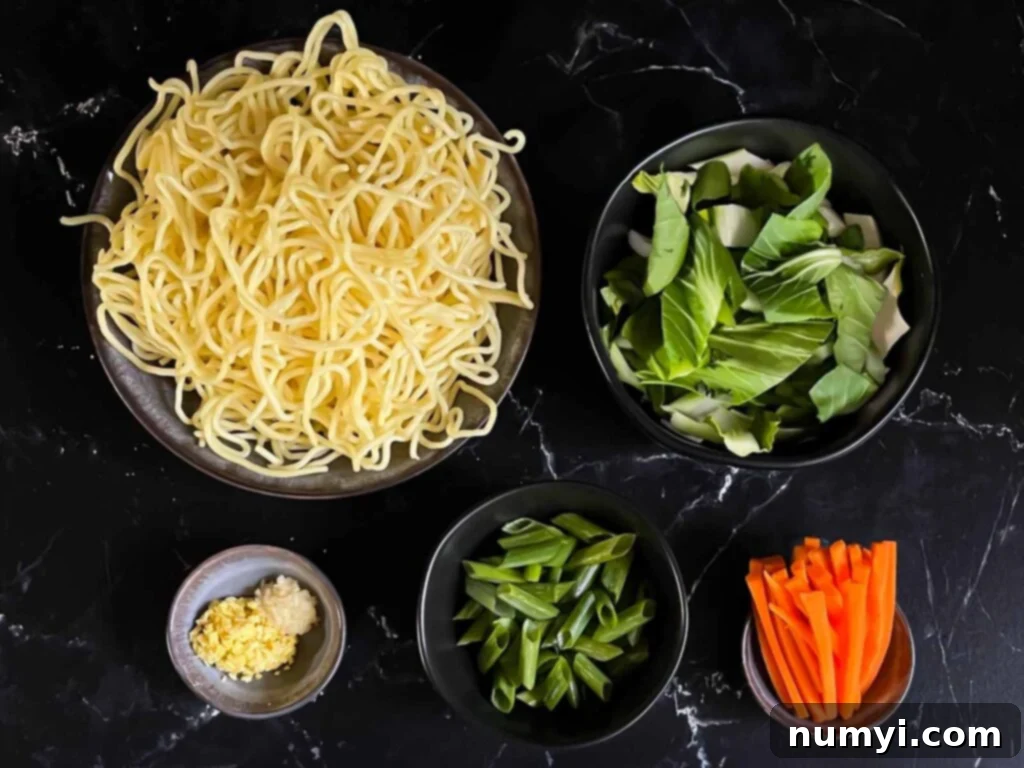 Finely diced ginger and garlic, along with sliced bok choy and carrots, meticulously prepped and ready for cooking a fresh vegetable lo mein dish, all arranged on a dark cutting board.