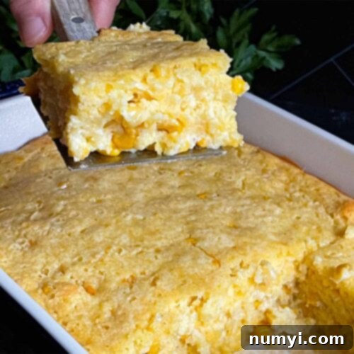 A serving of creamy corn casserole lifted over the baking pan on a dark surface, ready to be enjoyed.