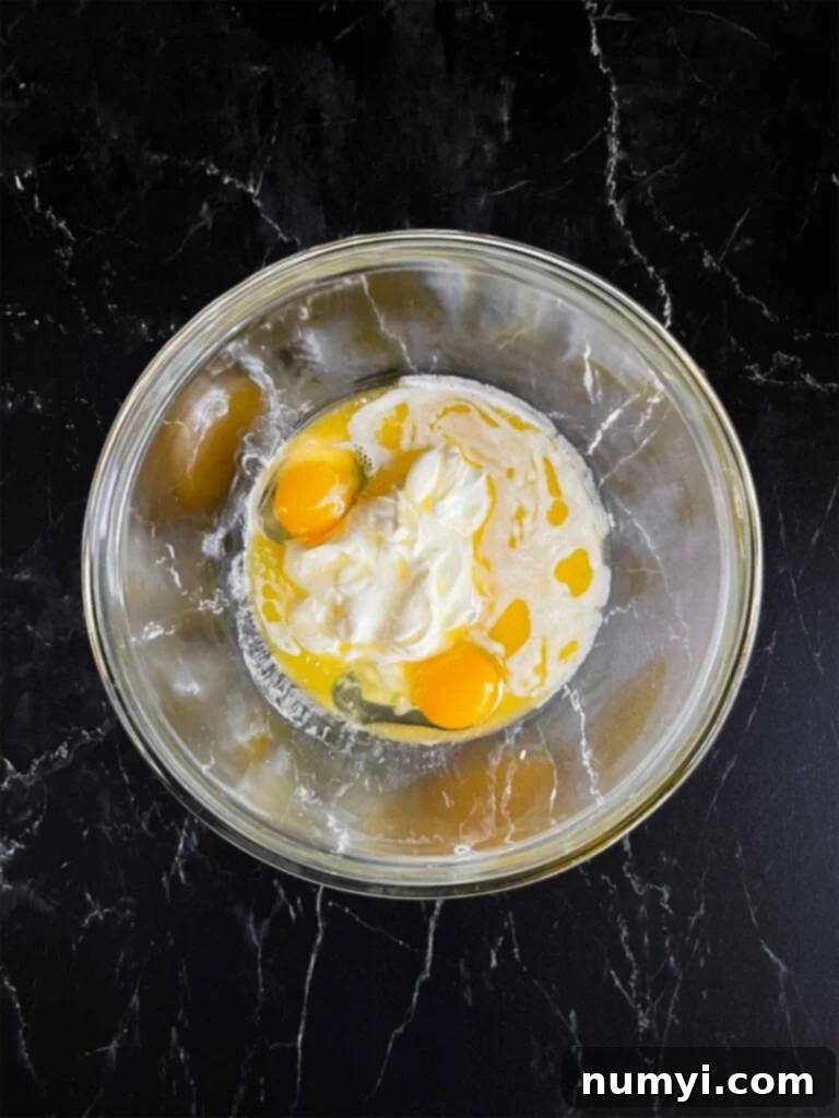 Wet ingredients for corn casserole, including eggs, sour cream, and melted butter, unmixed in a clear glass bowl.