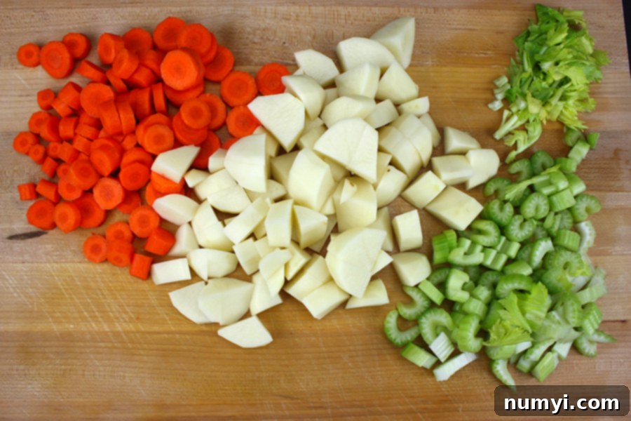 Assorted freshly diced carrots, potatoes, and celery with vibrant green leaves neatly arranged on a rustic wooden cutting board.