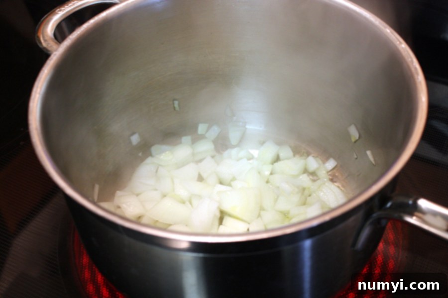 Finely chopped onions are being sautéed in golden olive oil within a large, heavy-bottomed metal pot, releasing a fragrant aroma.