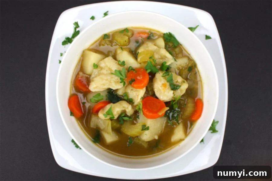 A beautifully presented bowl of vegetable dumpling soup on a crisp white square plate, elegantly garnished with a fresh sprinkle of chopped parsley, ready to be enjoyed.