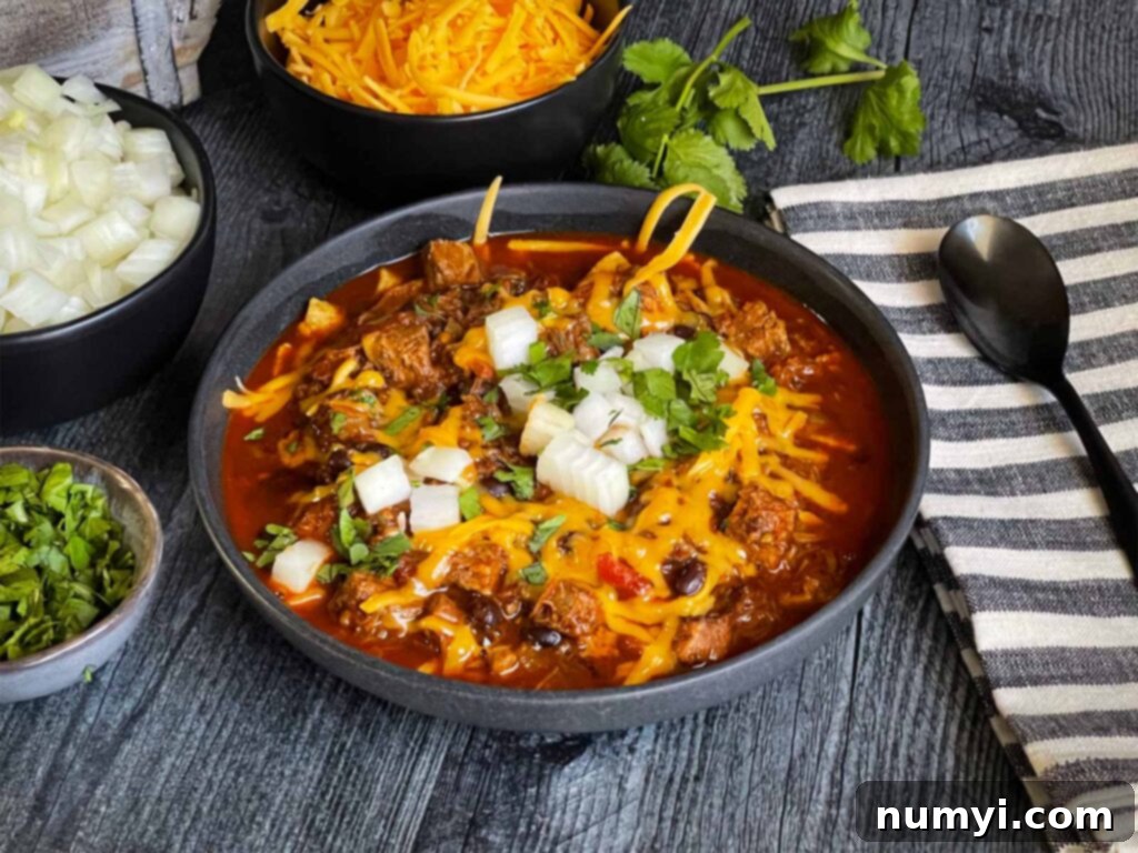 A hearty bowl of Smoked Texas Brisket Chili, generously garnished with fresh cilantro, diced white onions, and shredded cheddar cheese, resting on a rustic wooden surface. Steam gently rises from the chili, highlighting its warmth and inviting aroma.