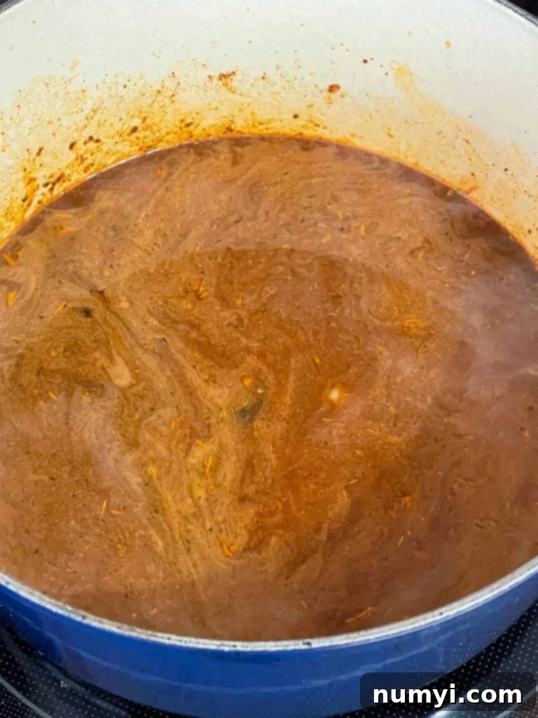 Cubed smoked brisket, beef broth, and black coffee mixed with the onion and spice base, simmering in a dark blue Dutch oven, forming the initial liquid phase of the chili.