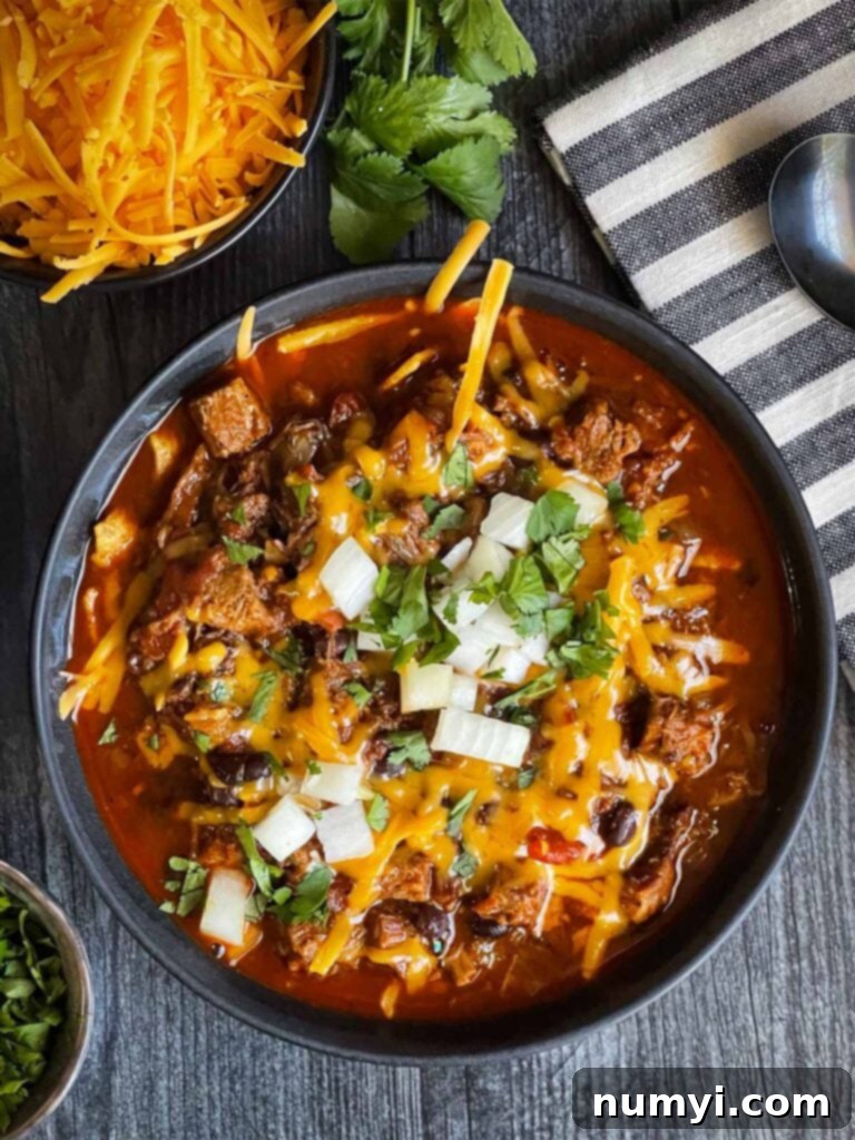 A vibrant bowl of Smoked Brisket Chili, artfully garnished with a sprinkle of fresh cilantro, finely diced white onions, and a generous layer of shredded cheddar cheese, presented on a natural wooden background.