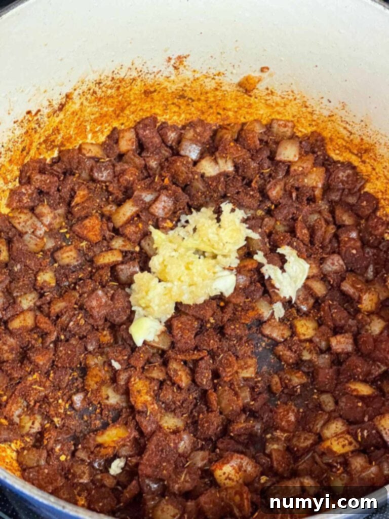 Freshly pressed garlic cloves being added to the cooked onions and spices in a Dutch oven, preparing the aromatic base for the brisket chili.