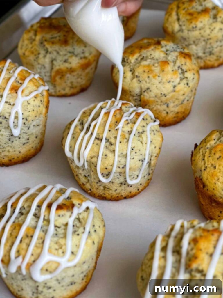 Lemon glaze being drizzled over the lemon poppy seed muffins on a parchment paper lined baking sheet.