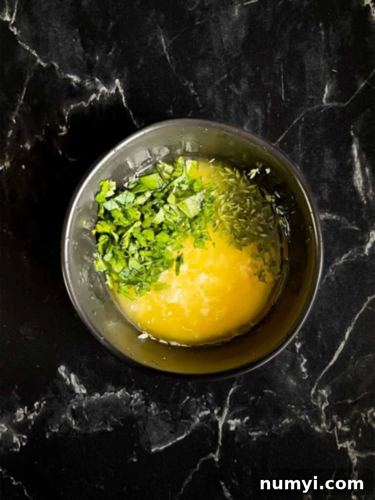 A dark bowl holding the garlic butter mixture, with melted butter, minced garlic, thyme, and parsley, on a dark surface.