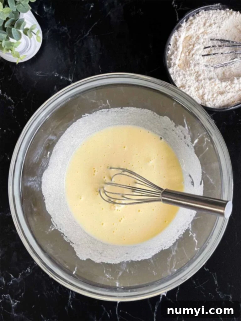 Eggs and granulated sugar beating until pale and thick in a glass mixing bowl with the dry ingredients in a dark bowl on a dark surface.
