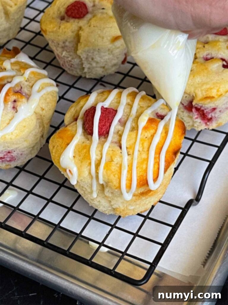 Lemon glaze being drizzled over the raspberry muffins on a wire rack over a parchment paper lined baking sheet.