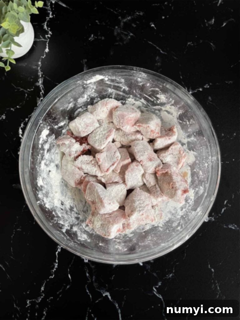 Seasoned beef tips dredged with flour in a glass mixing bowl on a dark surface.