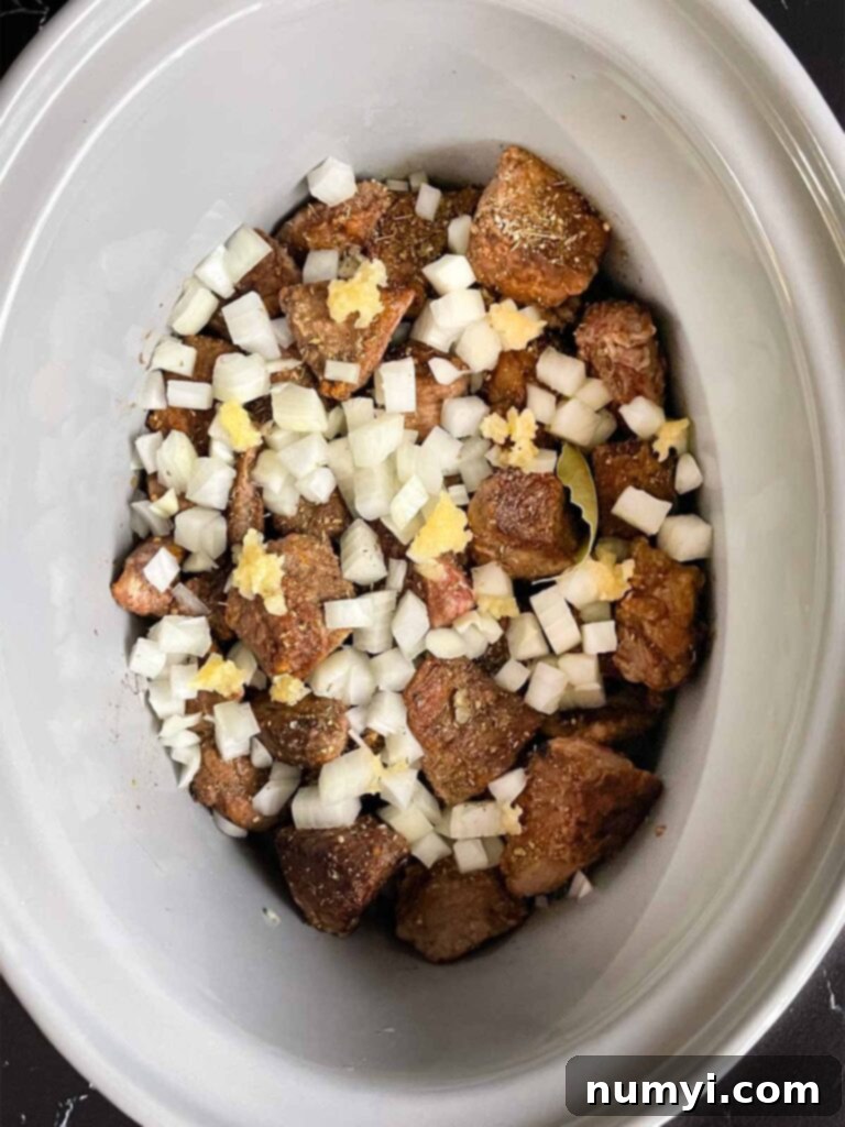 Seasonings, onions, and garlic added to the beef tips in the crock of a slow cooker.