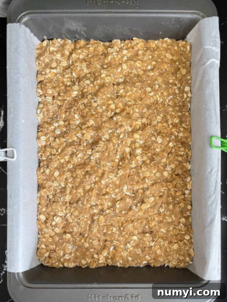 Oatmeal cookie dough for oatmeal fudge bars pressed into a baking pan lined with parchment paper.