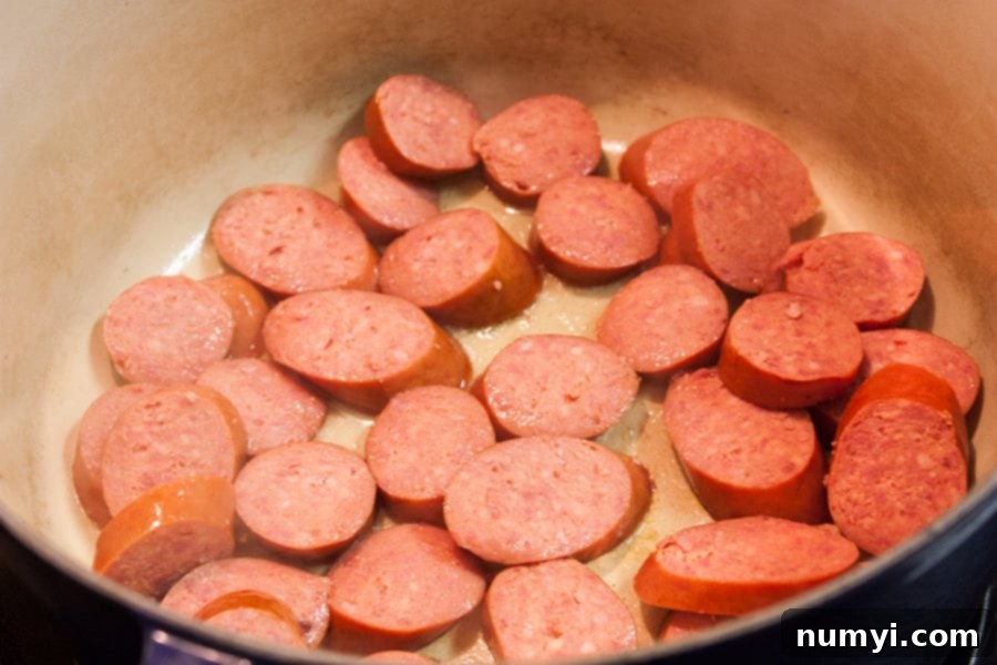 Andouille sausage slices browning in a large Dutch oven, releasing flavorful oils.