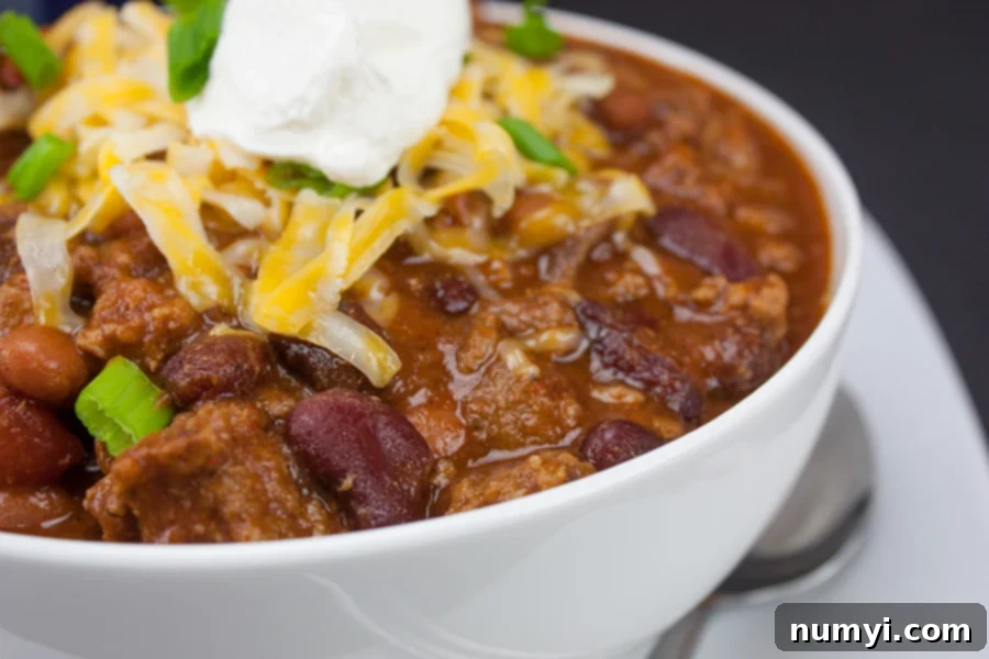 Thick and hearty homestyle chili with green onion, cheese, and sour cream as a garnish in a white bowl.