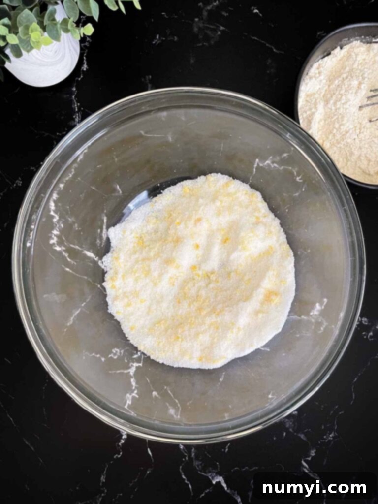 Granulated sugar and lemon zest rubbed together in a glass mixing bowl on a dark surface.