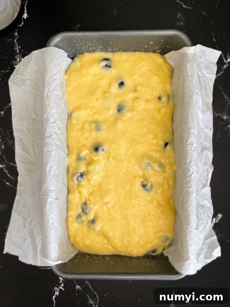Blueberry bread batter in a parchment paper lined loaf pan on a dark surface.