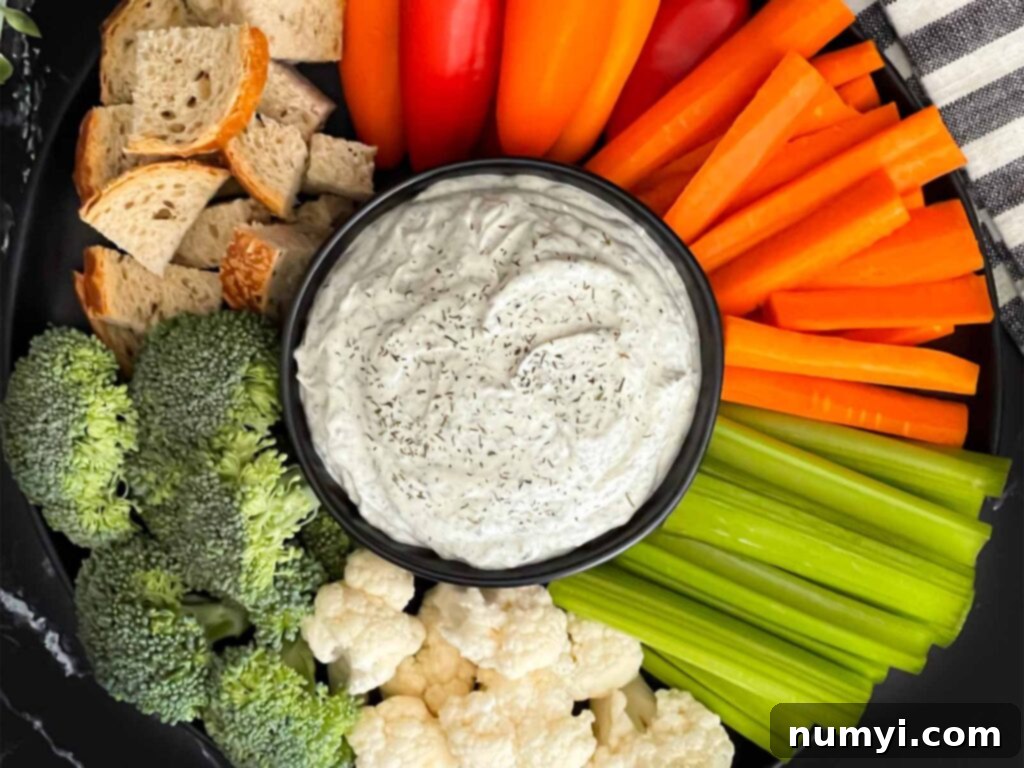 Dill dip in a small dark bowl surrounded by carrots, celery, cauliflower, broccoli, mini bell peppers and rye bread pieces on a dark plate.
