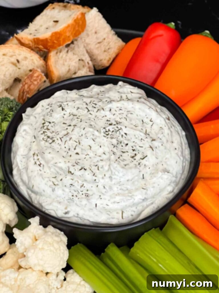 Dill dip in a small dark bowl surrounded by carrots, celery, cauliflower, broccoli, mini bell peppers and rye bread pieces on a dark plate.