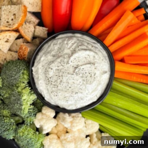 Dill dip in a small dark bowl surrounded by carrots, celery, cauliflower, broccoli, mini bell peppers and rye bread pieces on a dark plate.