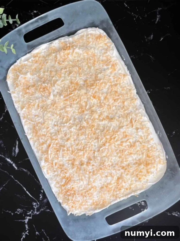 Lush Coconut Cream 10 Whipped topping spread over the coconut cream lush and sprinkled with toasted shredded coconut in a glass baking pan on a dark surface.
