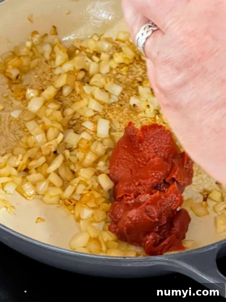 Tomato paste being added to a skillet with onions in it.