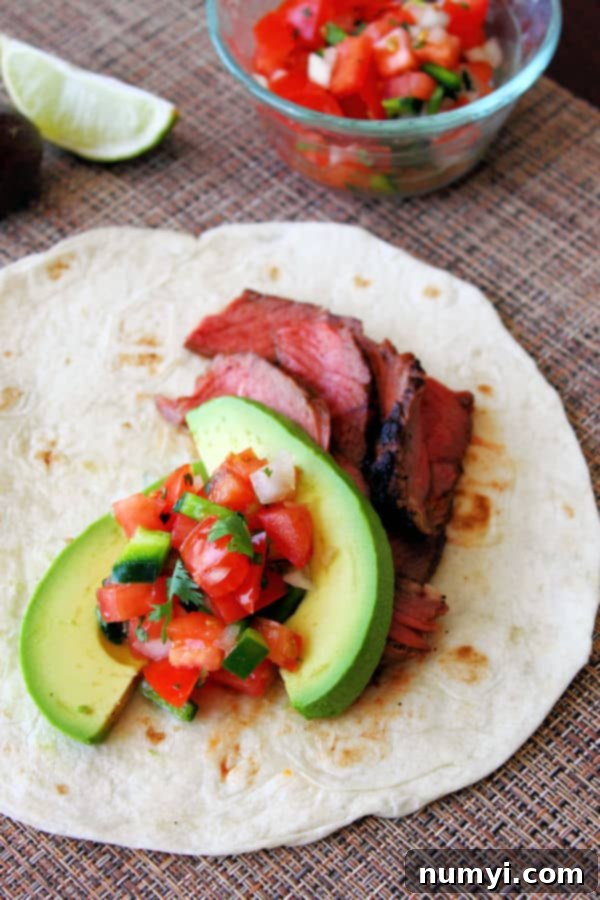 A close-up of a single grilled steak taco, perfectly assembled with a flour tortilla base, tender grilled steak strips, creamy avocado slices, and a colorful sprinkle of pico de gallo.