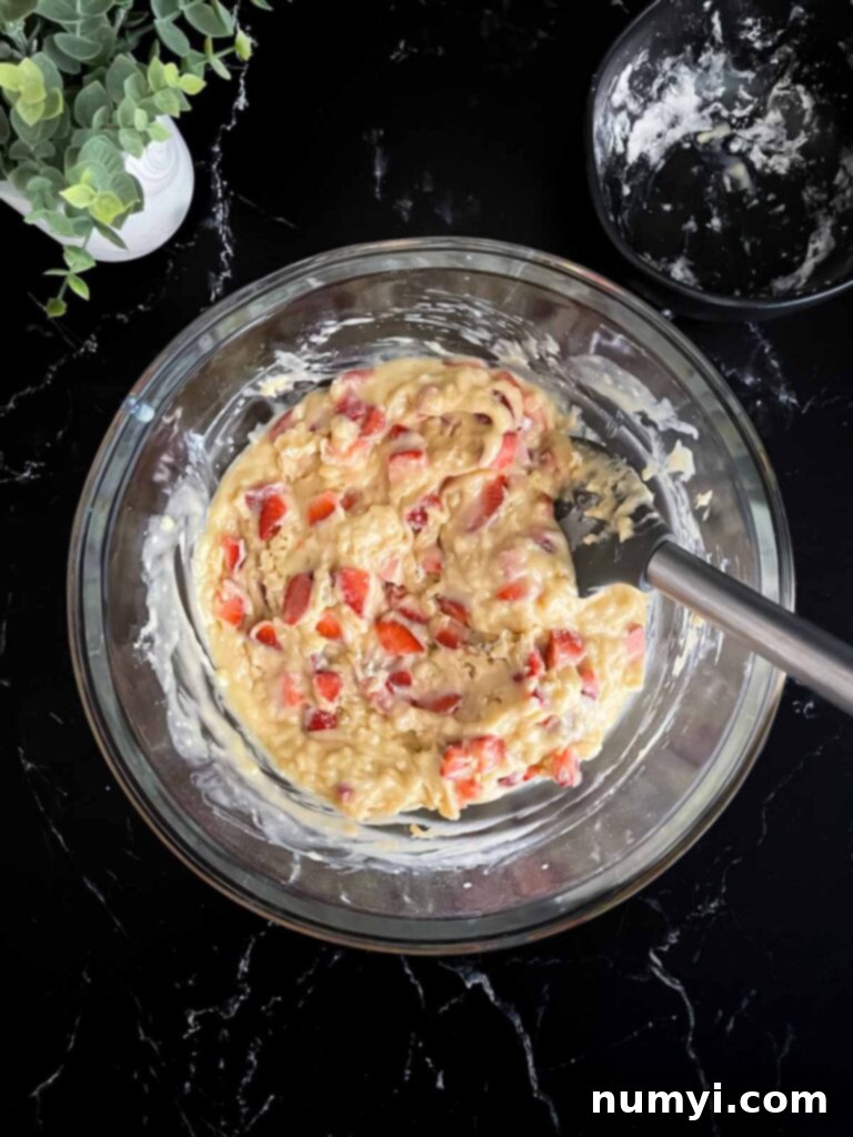 Strawberry muffin batter resting in a glass mixing bowl on a dark surface, covered with plastic wrap.