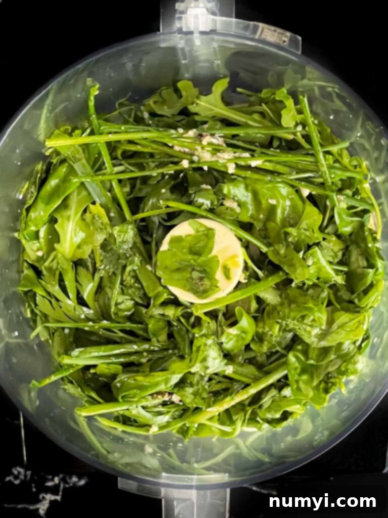 Fresh herbs, garlic, and liquids for the vinaigrette in the bowl of a food processor, ready to be blended.