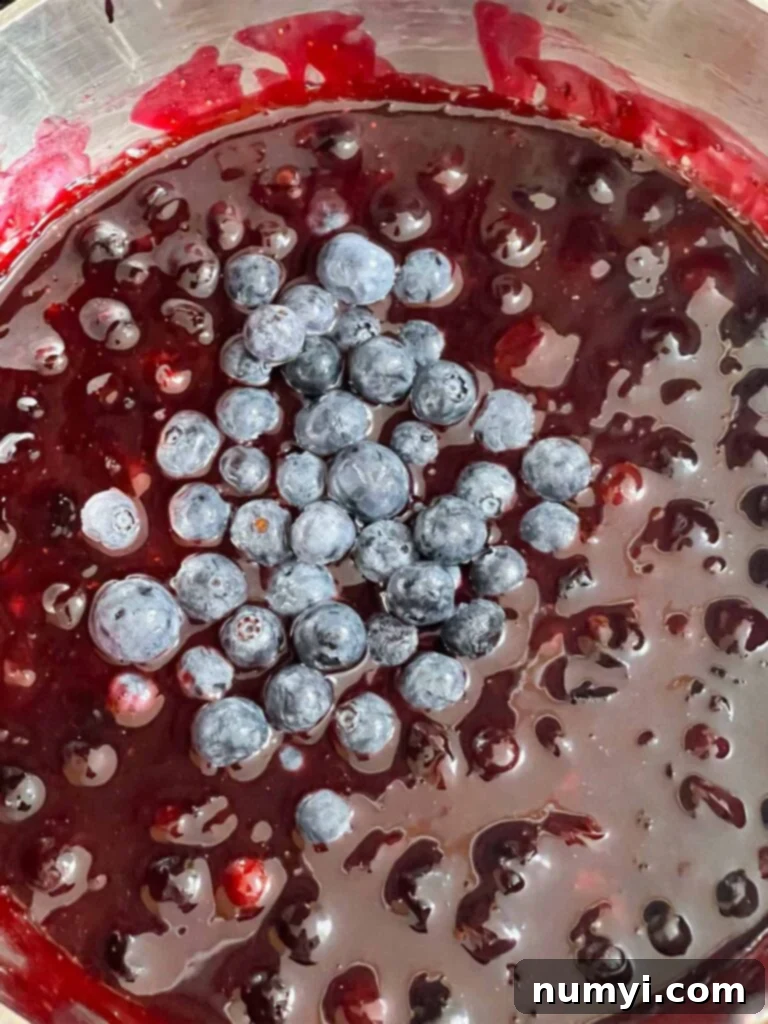 Luscious Berry Poke Cake 9 Ingredients for the blueberry filling, including fresh blueberries, lemon juice, sugar, and cornstarch, in a metal saucepan, simmering.