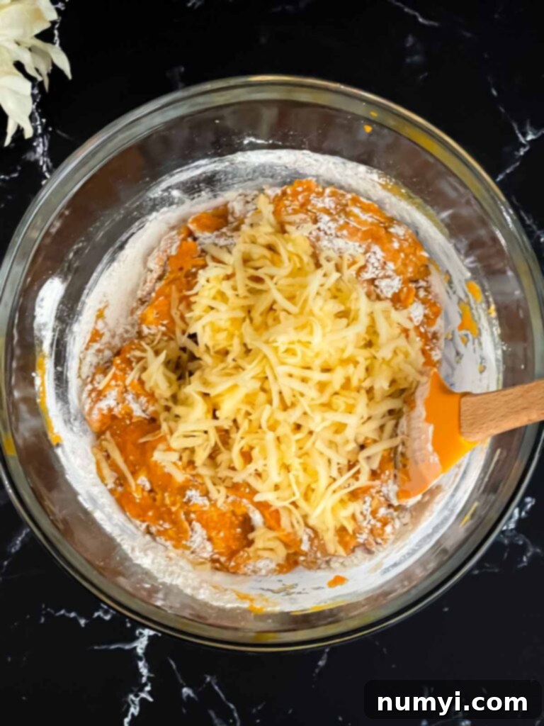 Shredded Honeycrisp apple being added to the rich, orange pumpkin muffin batter in a clear glass bowl, showcasing the moist ingredients.