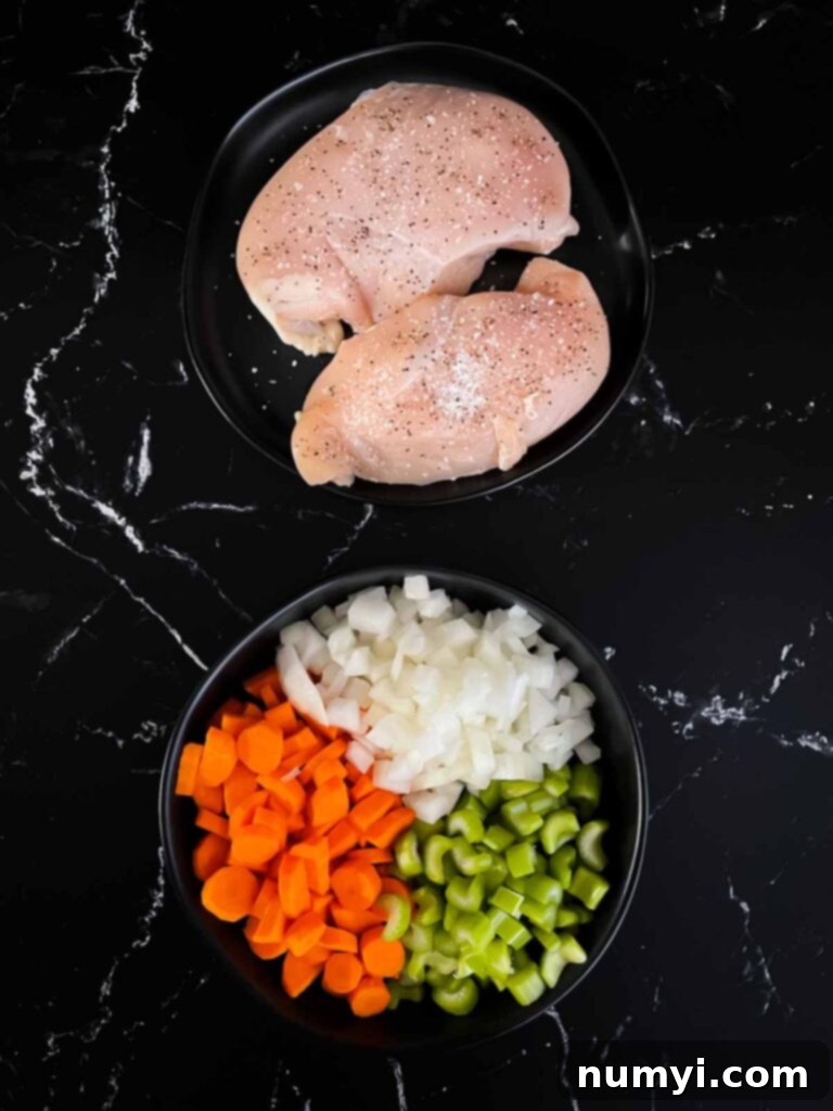 Chicken breasts on a dark plate seasoned with salt and pepper and a dark bowl full of diced onions, carrots, and celery.