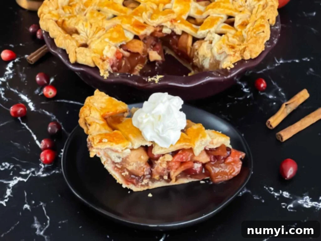 A perfectly baked slice of cranberry apple pie, adorned with a dollop of fresh whipped cream, resting on an elegant dark plate. The pie boasts a golden lattice crust, showcasing its rich, ruby-red filling.
