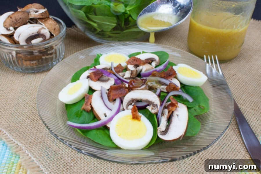 A vibrant spinach salad with crisp bacon, sliced mushrooms, hard-boiled eggs, and red onion, generously drizzled with a golden curry mustard vinaigrette, served on a clear glass plate.