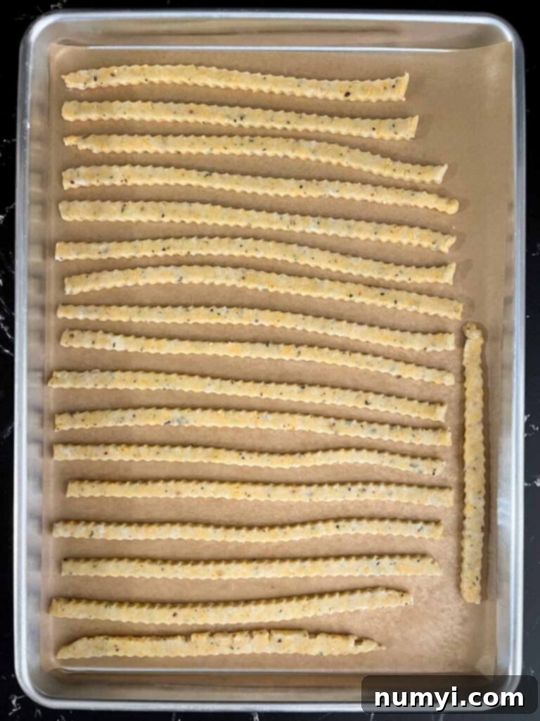 Crispy Cheddar Herb Sticks 12 Rows of raw cheddar cheese and herb straws meticulously arranged on a parchment-lined baking sheet, prepared for baking.