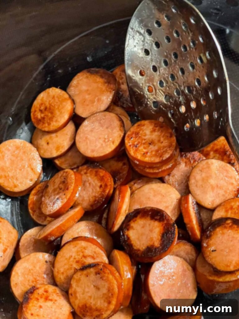 A spoon lifting browned smoked sausage slices from a Dutch oven, showing the crispy edges.
