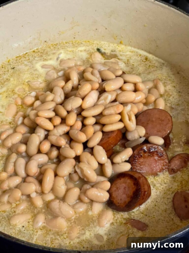 Adding canned Great Northern beans and browned smoked sausage back into the simmering soup in a Dutch oven.