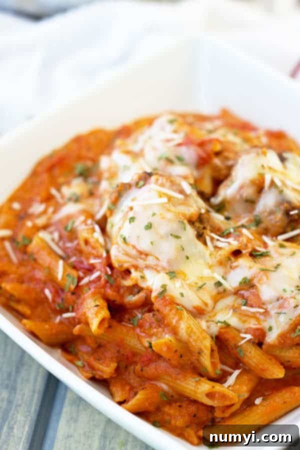 A close-up view of al dente penne pasta and perfectly cooked meatballs bathed in a rich, creamy marinara sauce, generously topped with melted mozzarella and grated Parmesan cheese.