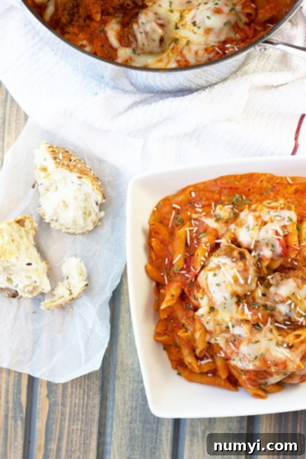 A perfectly portioned bowl overflowing with one-pot meatballs and penne pasta, garnished with a sprinkle of fresh herbs, placed next to several pieces of golden, crusty bread.