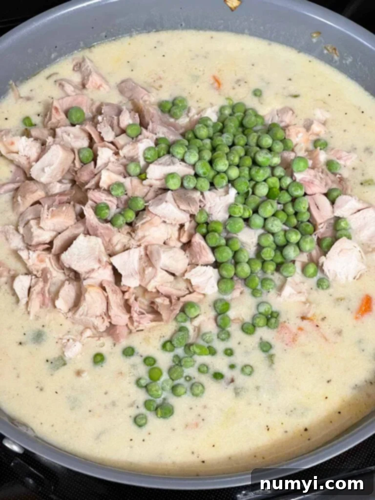 Adding cooked chicken and frozen peas to the creamy sauce mixture in a skillet.