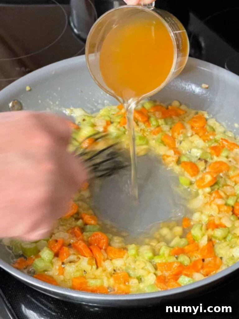 Pouring chicken stock into the skillet with mirepoix and roux, beginning to form the creamy sauce.